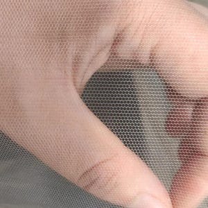 May include: A fine, semi-sheer, light beige mesh fabric featuring a consistent hexagonal pattern. The material is lightweight and translucent, displaying a delicate, grid-like structure that allows for visibility through the small, uniform openings.