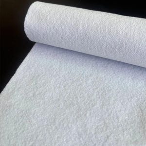 May include: A roll of white felt fabric featuring a dense, fibrous texture with a matte finish. The material is tightly wound into a cylindrical shape at the top, transitioning into a flat, smooth sheet of white textile that extends downward.