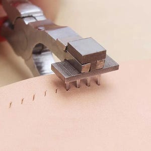 May include: A close-up of a leather sewing tool with a metal base and a set of prongs. The tool is being used to punch holes in a piece of leather.