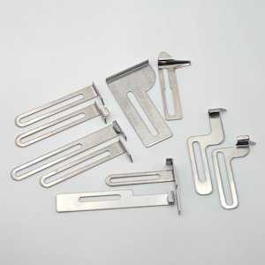 Può includere: Una collezione di varie staffe e clip metalliche in acciaio inossidabile, con diverse forme e dimensioni. Alcune hanno fessure allungate, mentre altre hanno design curvi o angolati. Il metallo ha una superficie lucida e riflettente.