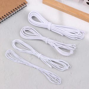 May include: Four bundles of white elastic cord, each tied in the middle. The cords are neatly arranged on a light gray surface, with a wooden element and a spiral-bound notebook visible in the background.