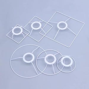 May include: A collection of six white metal lampshade frames in various sizes and shapes. Three frames are square with rounded corners, and three frames are circular. Each frame features a central ring supported by four thin metal spokes.