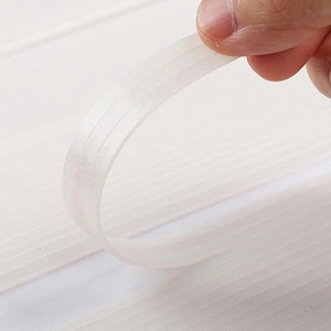 May include: A close-up of a white, semi-transparent ribbon being held up. The ribbon has subtle vertical lines and is slightly curved, creating a gentle arc. The background is a soft, white surface.