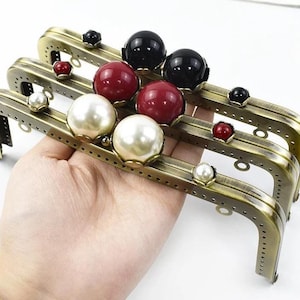 Può includere: Tre chiusure per borse in bronzo antico con decorazioni di perle e gioielli. Le cornici hanno una forma rettangolare con bordi arrotondati e sono decorate con grandi abbellimenti di perle e gioielli rossi, neri e bianchi.