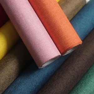 May include: Close-up of several rolls of colorful fabric. The rolls are in shades of pink, orange, blue, brown, and green. The fabric has a textured appearance, suggesting it may be linen or a similar material. The rolls are tightly wound.