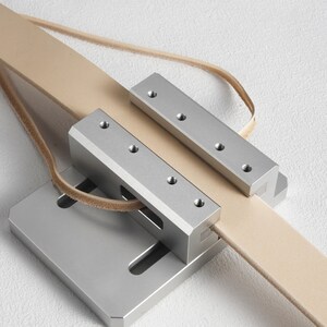 May include: A silver leather strop with a tan leather strap. The strop has a rectangular base and two metal bars with screw holes. The leather strap is looped through the metal components.