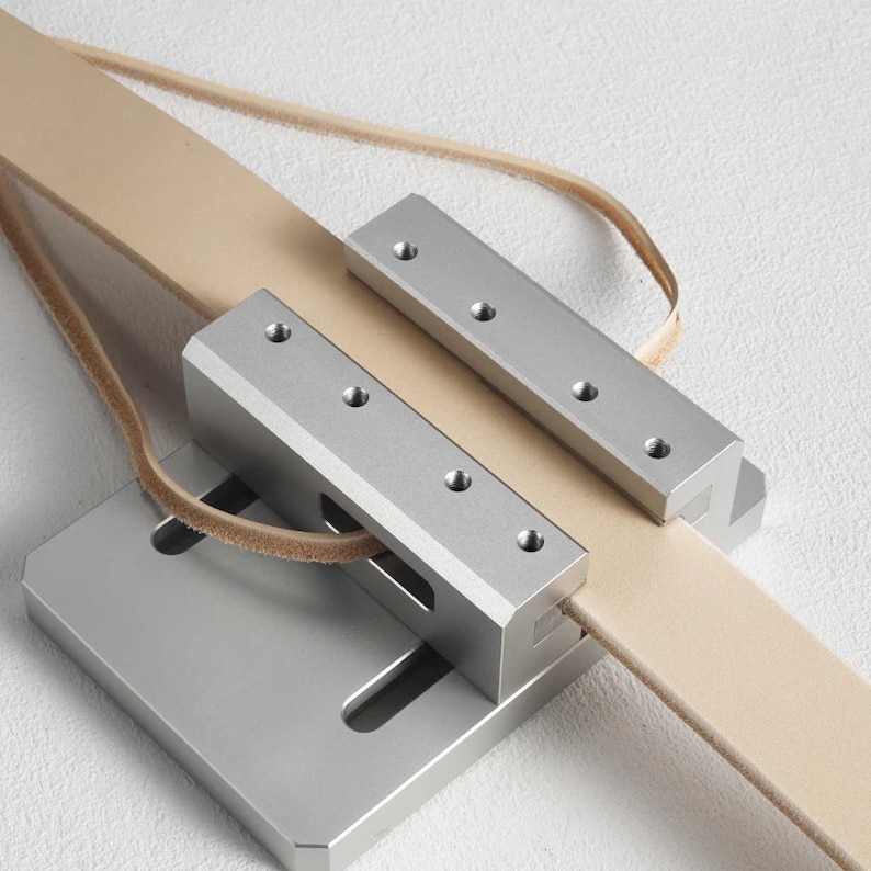 May include: A silver leather strop with a tan leather strap. The strop has a rectangular base and two metal bars with screw holes. The leather strap is looped through the metal components.