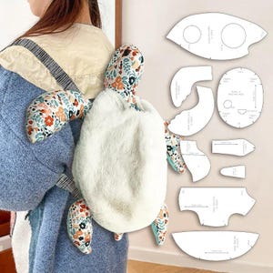 May include: A plush turtle backpack with a white, fluffy body and floral patterned flippers and head. The backpack is being worn by a person. Sewing patterns for the turtle are visible on the right side of the image.