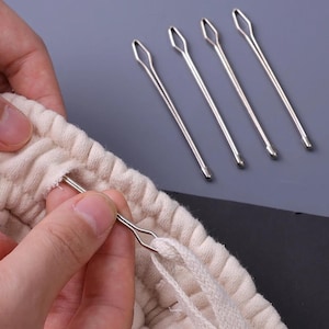 May include: Four silver-toned metal bodkins with elongated, diamond-shaped eyes for threading cord. One bodkin is inserted into the gathered waistband of a beige fabric garment, holding a white drawstring cord within its eye for easy threading.