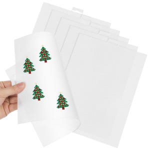 Könnte beinhalten: Ein Stapel weißer Kunststoff-Netzblätter für Handarbeiten, wobei das oberste Blatt vier kleine, gestickte grüne Weihnachtsbaum-Motive mit roten Ornamenten und einem gelben Stern an der Spitze im Kreuzstichmuster zeigt.
