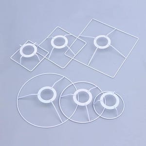 May include: A collection of white metal lamp shade frames in various shapes and sizes. The frames include square and circular designs, with a central ring and supporting wire spokes. The frames are arranged on a light gray surface.