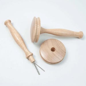 Op de afbeelding: Een set van drie houten gereedschappen voor het winden van garen. De gereedschappen zijn gemaakt van lichtbruin hout en hebben een gladde afwerking. De set bevat een garenwinder, een garenwals en een garenspeld.