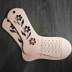 May include: Two light-coloured wooden sock blockers with a smooth finish. Each blocker features a cutout floral pattern along the ankle and a grid of small holes with printed numerical sizing indicators on the heel and foot area for knitting.