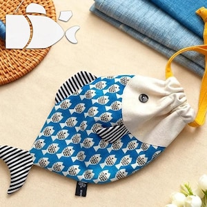 Puede incluir: Una bolsa con cordón azul en forma de pez con cabeza y cola blancas, y aletas a rayas blancas y negras. La bolsa tiene un estampado de peces blancos sobre un fondo azul. Se adjunta una correa amarilla.