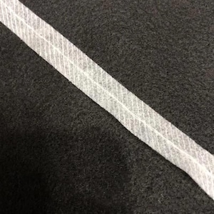 Puede incluir: Una tira de cinta adhesiva termoadhesiva blanca semitransparente con una línea de pliegue central y una textura fina de cuadrícula. La cinta está colocada en diagonal sobre una superficie de tela oscura con textura de color carbón.