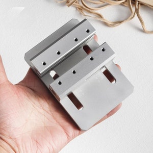 May include: A silver metal tool with multiple drilled holes and slots. The tool has a rectangular, layered design, held in a person's hand. The tool appears to be a precision instrument, possibly for woodworking or metalworking.