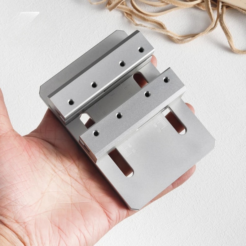 May include: A silver metal tool with multiple drilled holes and slots. The tool has a rectangular, layered design, held in a person's hand. The tool appears to be a precision instrument, possibly for woodworking or metalworking.
