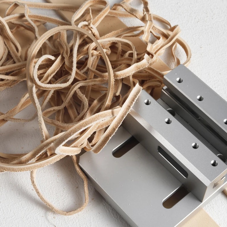 May include: A metal leather strip cutter with tan leather strips. The cutter is silver and has a rectangular base with two parallel blades. The leather strips are curled and have a textured appearance. The image is a close-up.