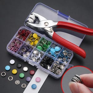 Peut inclure: Un organisateur en plastique transparent rempli de boutons-pression colorés et une pince à boutons-pression argentée et rouge. Les attaches sont de différentes couleurs, dont le rouge, le jaune, le vert, le bleu et le noir. Une bande de tissu blanc est également incluse.