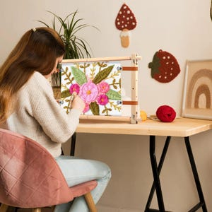 May include: A person working on a floral embroidery project. The embroidery features pink and green flowers on a white background, held in a wooden frame. A red ball of yarn and other craft supplies are on the table.