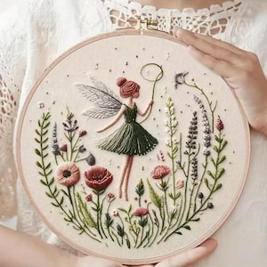 May include: Embroidered fairy hoop art featuring a fairy in a green dress with wings, surrounded by colorful flowers and greenery. The fairy is holding a loop. The embroidery is set in a wooden hoop.