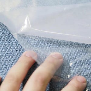 Può includere: Primo piano di una mano che tocca un foglio di plastica trasparente e trasparente. La plastica è piegata, creando un angolo acuto. Lo sfondo è un tessuto blu strutturato.