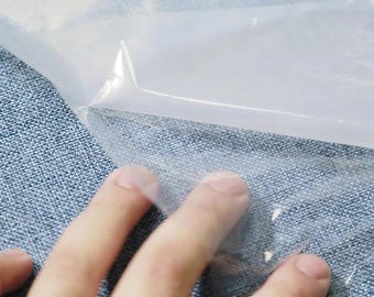 May include: A close-up of a hand touching a clear, transparent plastic sheet. The plastic is folded over, creating a sharp angle. The background is a textured blue fabric.