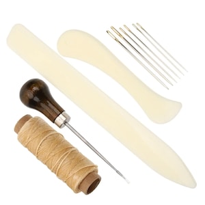 Puede incluir: Un kit de artesanía en cuero con un punzón con mango de madera, un carrete de hilo beige, varias agujas y plegaderas. Las herramientas están dispuestas sobre un fondo blanco, ideal para proyectos de marroquinería.