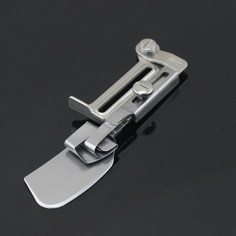 K&ouml;nnte beinhalten: Ein silberfarbener N&auml;hmaschinen-Binderaufsatz. Der Aufsatz hat eine flache, abgewinkelte Basis mit einer gebogenen Kante, einen Klappclip und verstellbare Schrauben. Das Metallwerkzeug dient zum Falten von Stoffkanten zum N&auml;hen.
