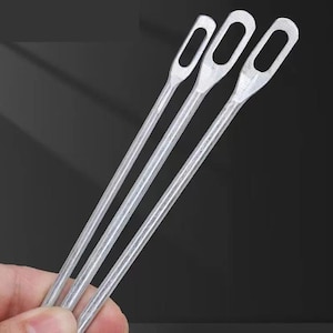 Puede incluir: Tres herramientas de metal plateado con aberturas ovales alargadas en la parte superior. Las herramientas tienen mangos largos, delgados y rectos. Las herramientas se sostienen en la mano de una persona contra un fondo oscuro.