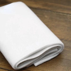 Peut inclure: Une pièce de feutre blanc uni pliée avec une texture douce et mate. Le matériau est opaque et présente une couleur uniforme et cohérente, affichant une surface propre et lisse avec des couches visibles là où le tissu est replié sur lui-même.