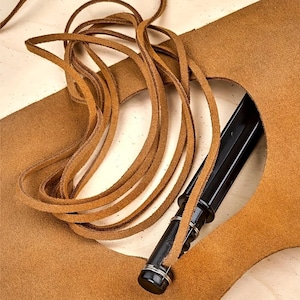 Könnte beinhalten: Nahaufnahme eines Lederhandwerksprojekts. Das Bild zeigt ein schwarzes Messer mit einer braunen Lederscheide und langen, dünnen Lederstreifen. Das Leder hat eine warme braune Farbe, und der Messergriff hat silberne Akzente.