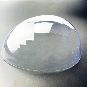 May include: A clear, transparent, dome-shaped object with a smooth, curved surface. The item has a rounded, semi-spherical form that is hollow and open at the bottom, featuring a polished, glass-like appearance with subtle light reflections.