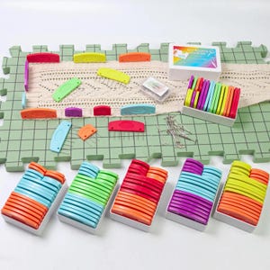 Può includere: Un set di marcatori per punti a maglia colorati in varie forme e dimensioni, organizzati in contenitori di plastica trasparente. I marcatori sono disposti in file di diversi colori, tra cui arancione, rosso, verde, blu e viola. Il set include anche un piccolo contenitore di ferri da maglia.