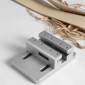 May include: A silver metal leathercraft tool with a rectangular base and two parallel, raised sections. The tool has several small holes and slots. Light brown leather strips are in the background.