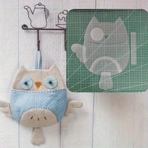 May include: A beige and light blue polka dot fabric owl ornament with two round eyes and a small orange beak. Beside it, a transparent plastic owl-shaped sewing template rests on a green cutting mat with white grid lines and diagonal markings.