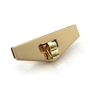 May include: A gold-coloured metal clasp with a geometric design. The clasp has a rectangular body with a beveled edge and a small, rounded loop. The loop is attached to the body with a hinge. The clasp is set against a white background.