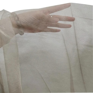 May include: A sheer, light beige fabric is held up, allowing light to pass through. The fabric appears to be a delicate material, possibly tulle or chiffon, with a subtle texture. The image highlights the fabric's transparency and drape.