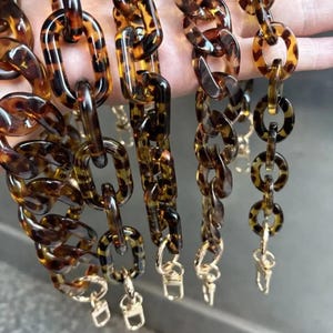 Puede incluir: Una colección de eslabones de cadena acrílicos con estampado de carey marrón y beige. Los eslabones están conectados con cierres dorados. Las cadenas están diseñadas para usarse como correa de bolso o accesorio.