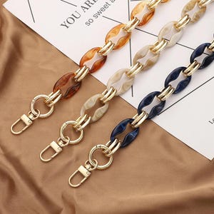 Op de afbeelding: Drie kleurrijke portemonnee kettingen met goudkleurige hardware. De kettingen hebben ovale schakels in bruin, crème en marineblauw. Elke ketting heeft een karabijnhaak voor eenvoudige bevestiging aan een tas. De achtergrond bevat de tekst "YOU ARE so sweet a".