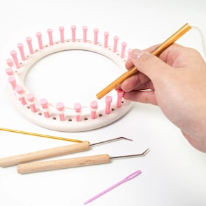 May include: A white circular knitting loom with pink pegs is shown, along with a wooden crochet hook and two wooden-handled crochet tools. A hand is using the crochet hook on the loom. A pink plastic yarn needle is also present.