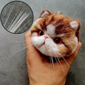 Puede incluir: Adorno artesanal de cabeza de gato con un patrón marrón y blanco, ojos rojos y largos bigotes blancos. El adorno se sostiene en una mano, mostrando la artesanía detallada. Un primer plano muestra los mechones de bigotes individuales.