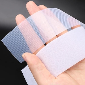 May include: Close-up of a hand holding strips of hook and loop fastener. The image shows a white, fuzzy strip and a clear, textured strip with a blue and pink hue. The background is a dark, solid color.