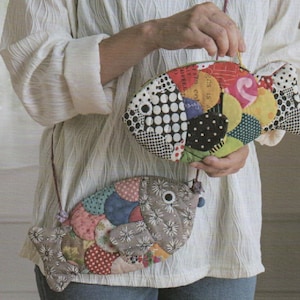 May include: Two fish-shaped purses with long straps. The purses are made of patchwork fabric in various colors and patterns, including polka dots, floral prints, and solid colors. The purses have a zipper closure and a small loop for carrying.
