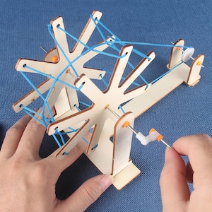 May include: A wooden toy with a spinning wheel and blue string. The wheel is connected to a crank that can be turned to make the wheel spin. The string is wrapped around the wheel and is used to create a pattern.