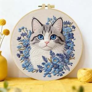 May include: A circular embroidery hoop containing a fabric panel with a detailed needlework portrait of a grey and white cat with bright blue eyes, surrounded by a dense arrangement of small, vibrant blue flowers and green foliage.