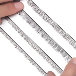 Könnte beinhalten: Nahaufnahme von mehreren Streifen falscher Wimpern. Jeder Streifen ist schwarz und weist einzelne, feine, dunkle Wimpern auf. Die Streifen sind diagonal vor einem weißen Hintergrund angeordnet, mit Fingern, die die Kanten halten.