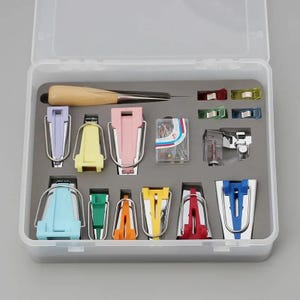 Op de afbeelding: Een transparante plastic opbergdoos gevuld met kleurrijke naai-gereedschappen, waaronder naaldverwijderaars, clips, spelden en een houten priem. De doos is georganiseerd met vakken voor elk gereedschap.