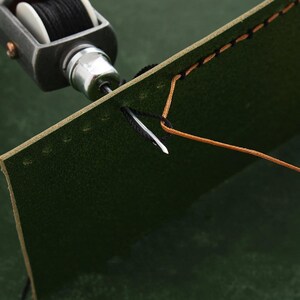 May include: A close-up of a leather sewing awl with a needle threading through a piece of green leather. The leather is stitched with black and brown thread.