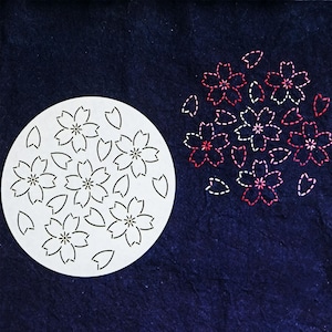 Puede incluir: Un esténcil circular blanco con diseños de flores de cerezo recortadas y formas de hojas. A la derecha, flores de cerezo bordadas en hilo blanco y rojo se muestran sobre un fondo de tela azul oscuro. El diseño evoca la primavera y la naturaleza.